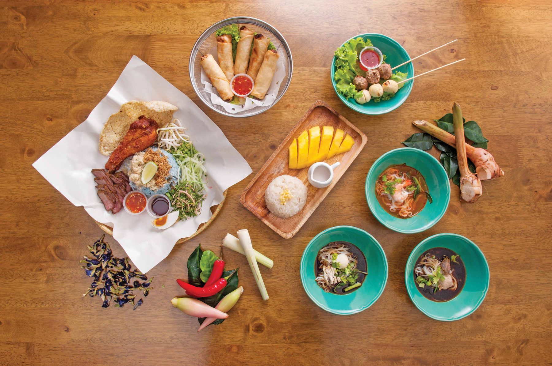 A full table at Thamada: boat noodle in teal bowls, nasi kerabu, khao kluk kapi, spring rolls, mango sticky rice